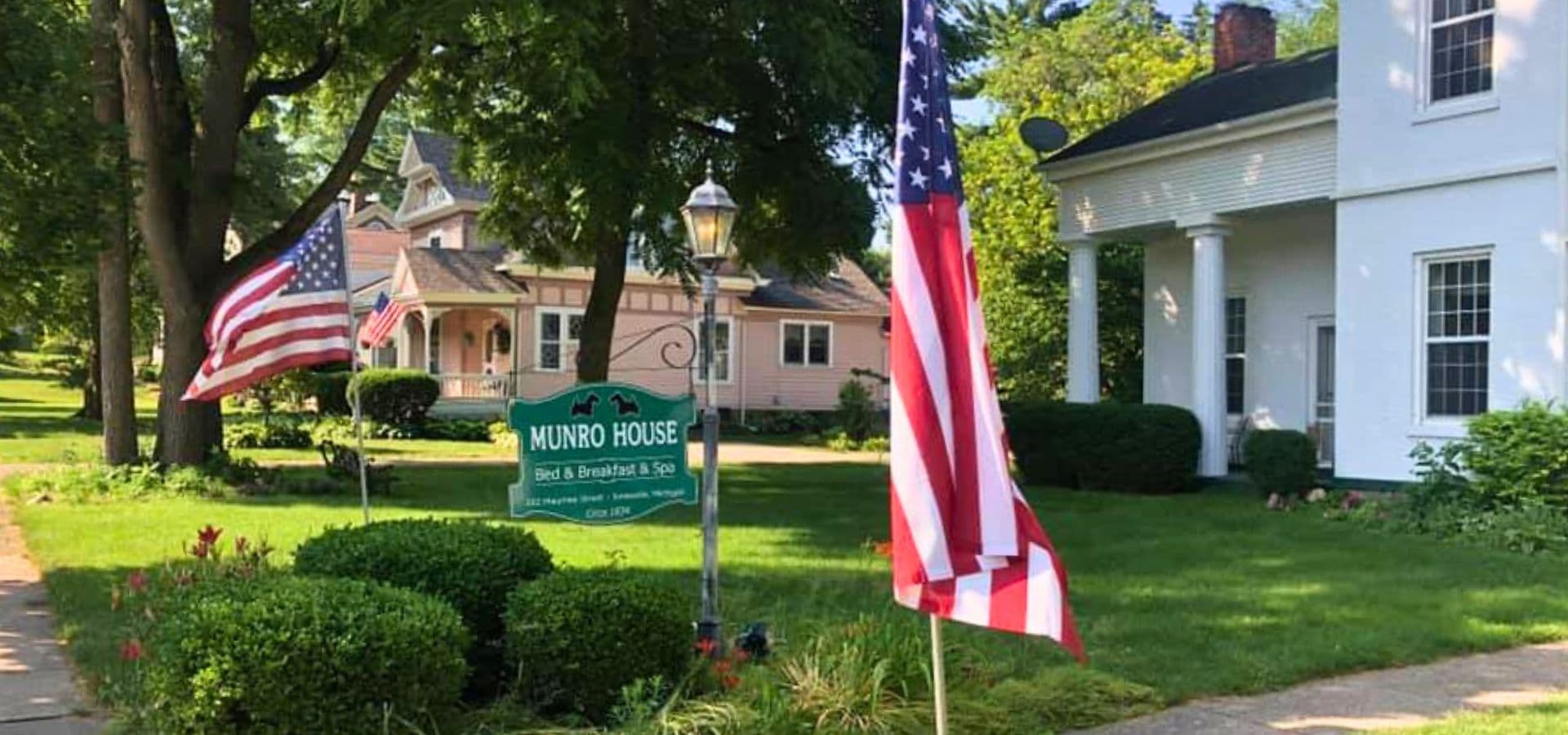Two American flags stand in front of the Munro House bed and breakfast, surrounded by greenery.
