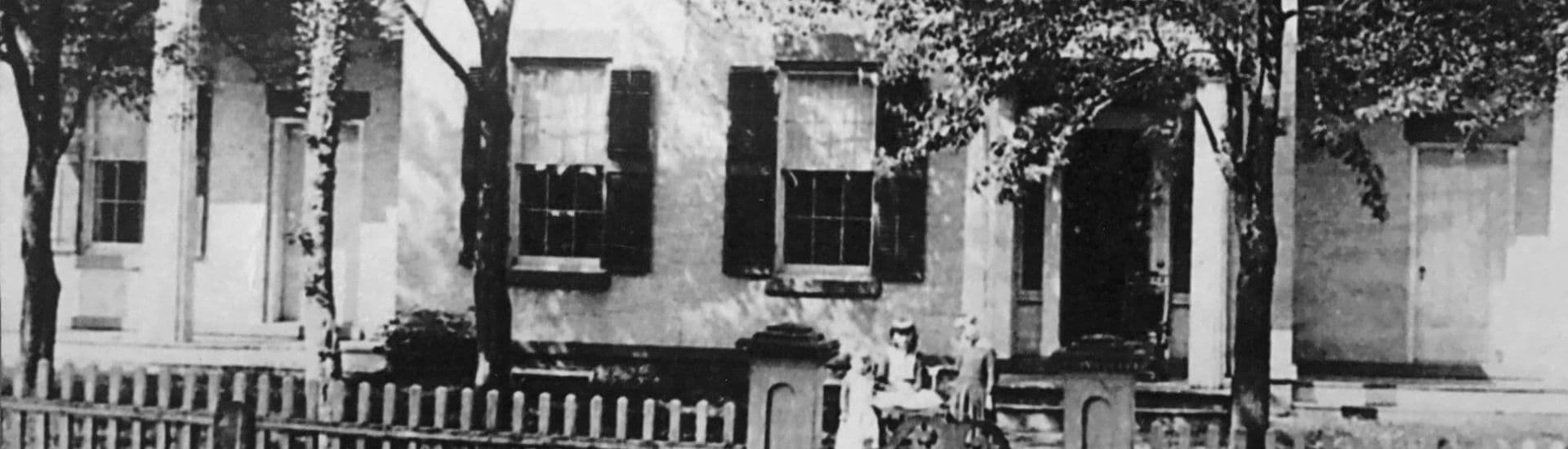 A historical black-and-white photo of a modest house with trees and a picket fence, featuring children seated in front.
