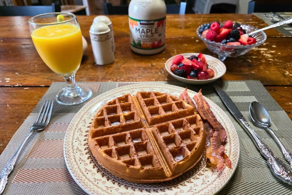 A breakfast plate featuring a waffle, bacon, a fruit bowl, and a glass of orange juice.