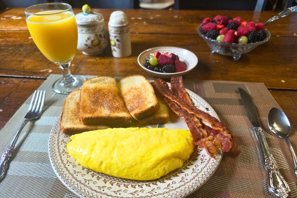 A breakfast plate featuring an omelet, toast, bacon, a glass of orange juice, and a bowl of mixed berries.