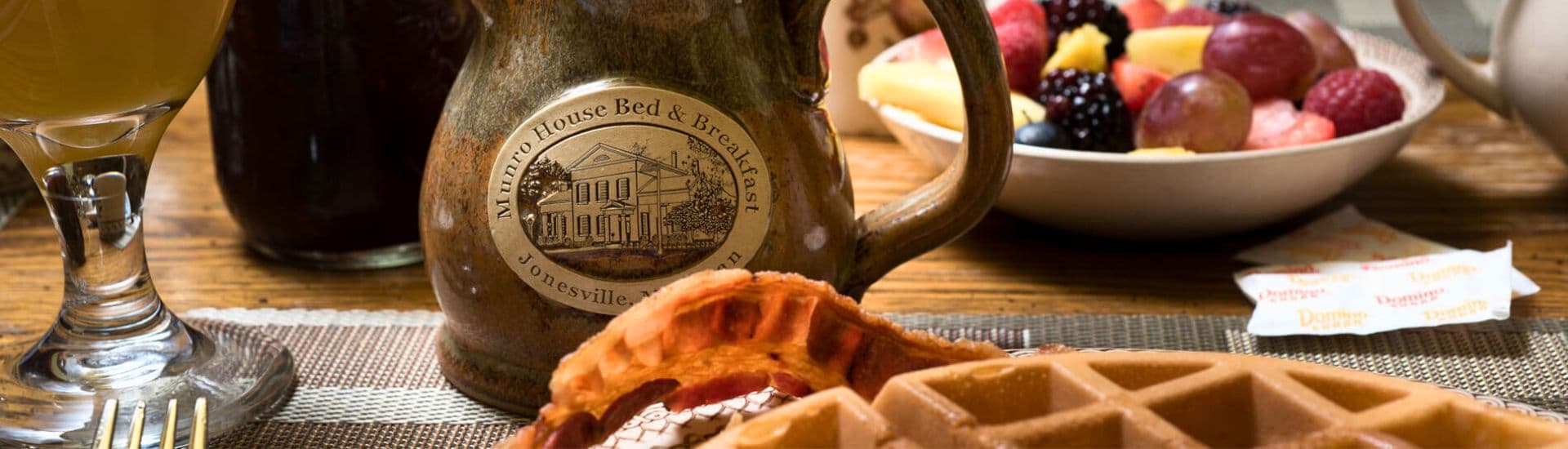 A breakfast table featuring a decorative pitcher, waffles, bacon, and a bowl of assorted fruits.