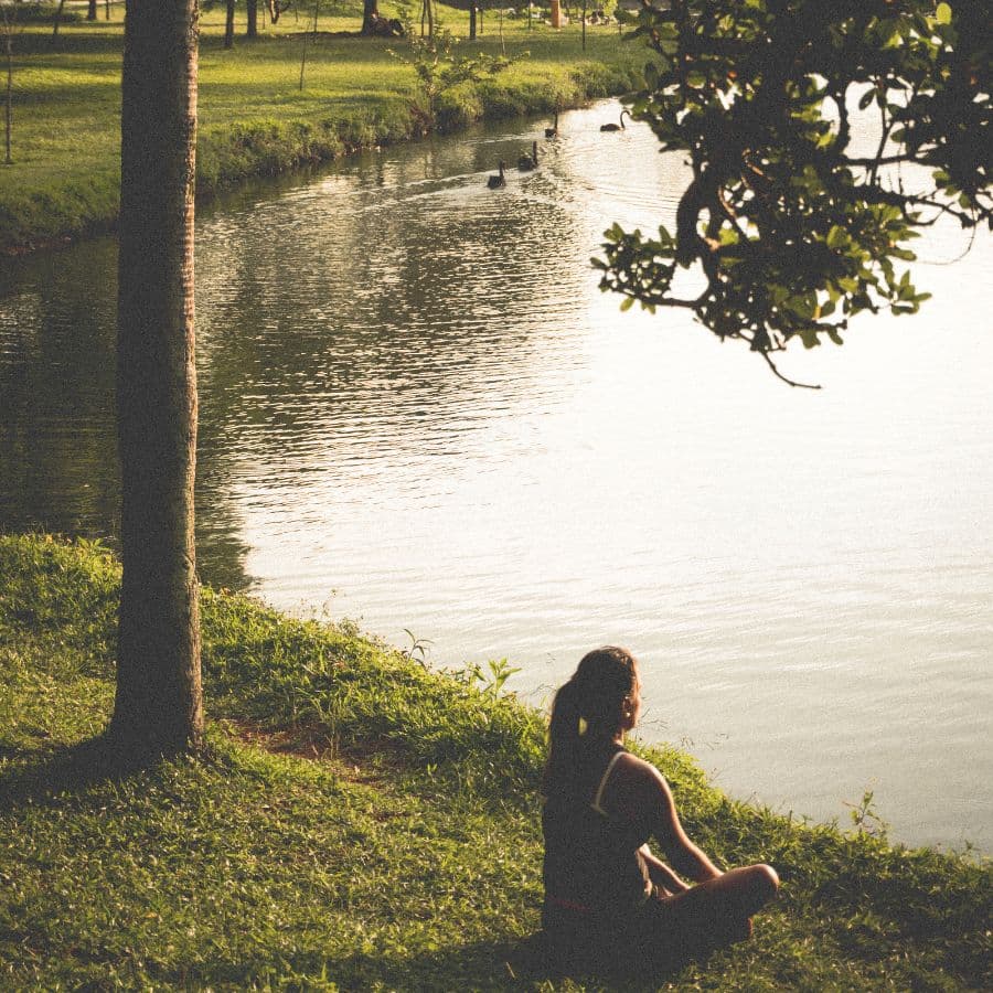 A person sits by a tranquil lake surrounded by greenery.