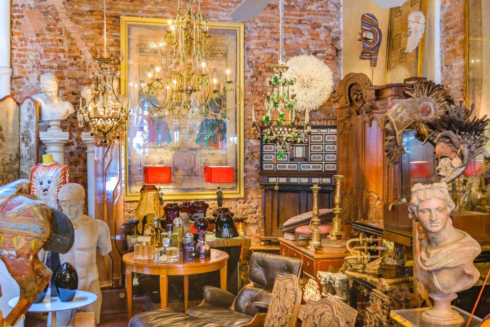 A vibrant antique shop filled with sculptures, ornate chandeliers, and eclectic decorative items against a brick wall.