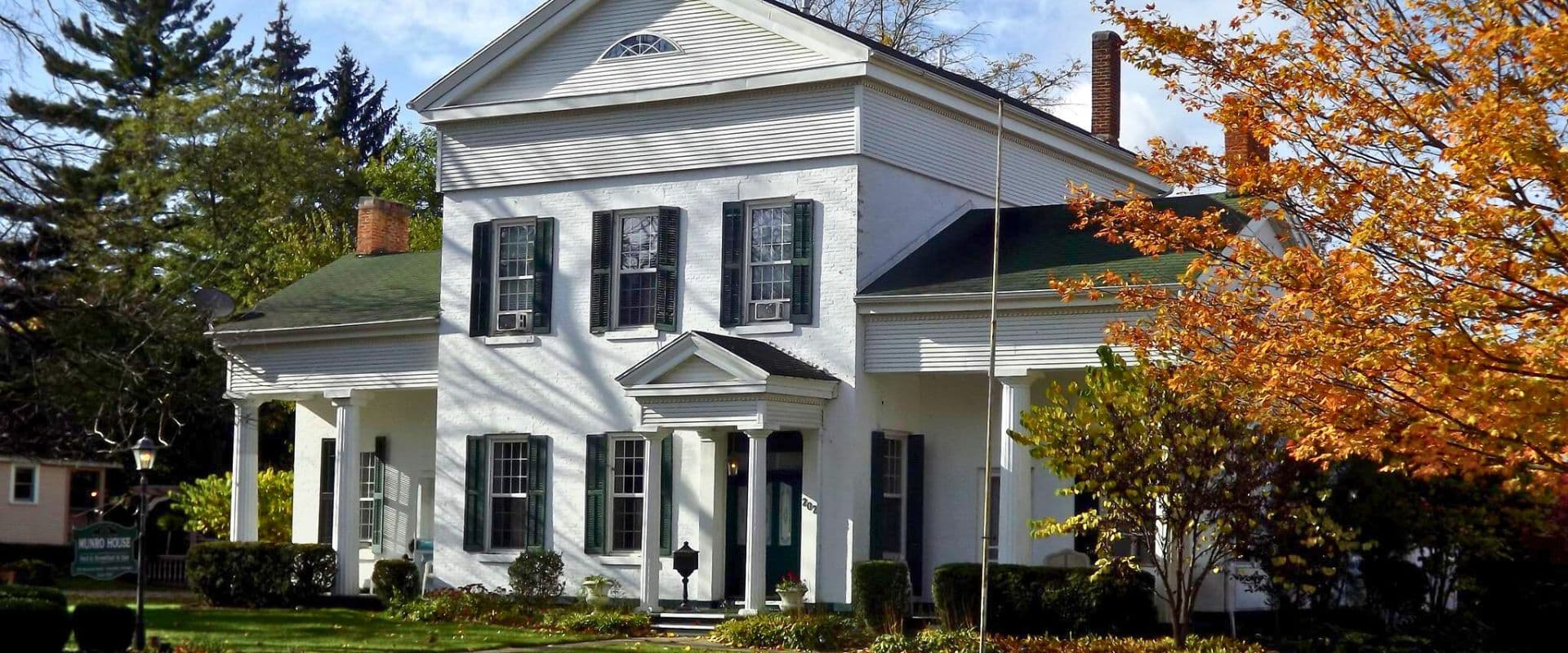 A classic white house with green shutters and a sloped roof, surrounded by trees and autumn foliage.