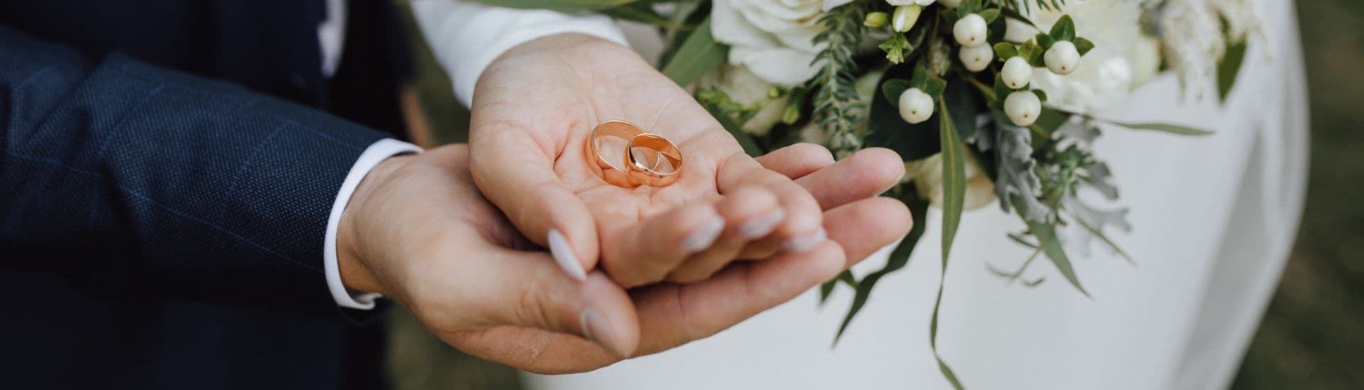 A hand holds two wedding rings with a bouquet in the background.