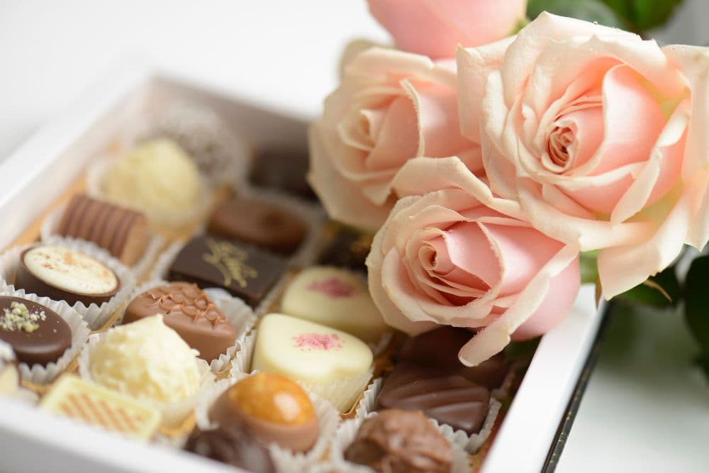 A box of assorted chocolates beside pale pink roses.