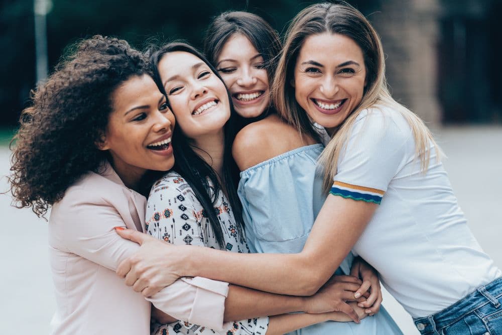 Four smiling women joyfully embracing each other outdoors.