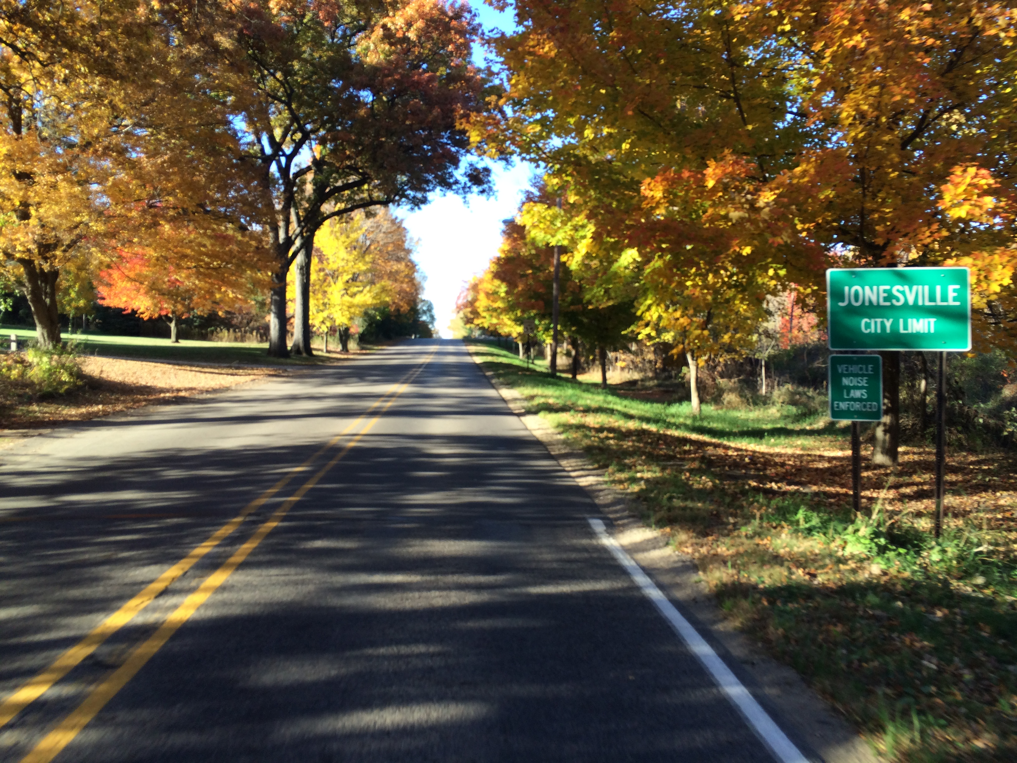 Blacktop road with yellow centerline surrounded by trees in brilliant fall colors of yellow and orange with a green Jonesville City Limit sign