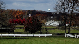 Horse farm with white fence before grey barn at the edge of a forest of Fall colors with a white water tower in the background