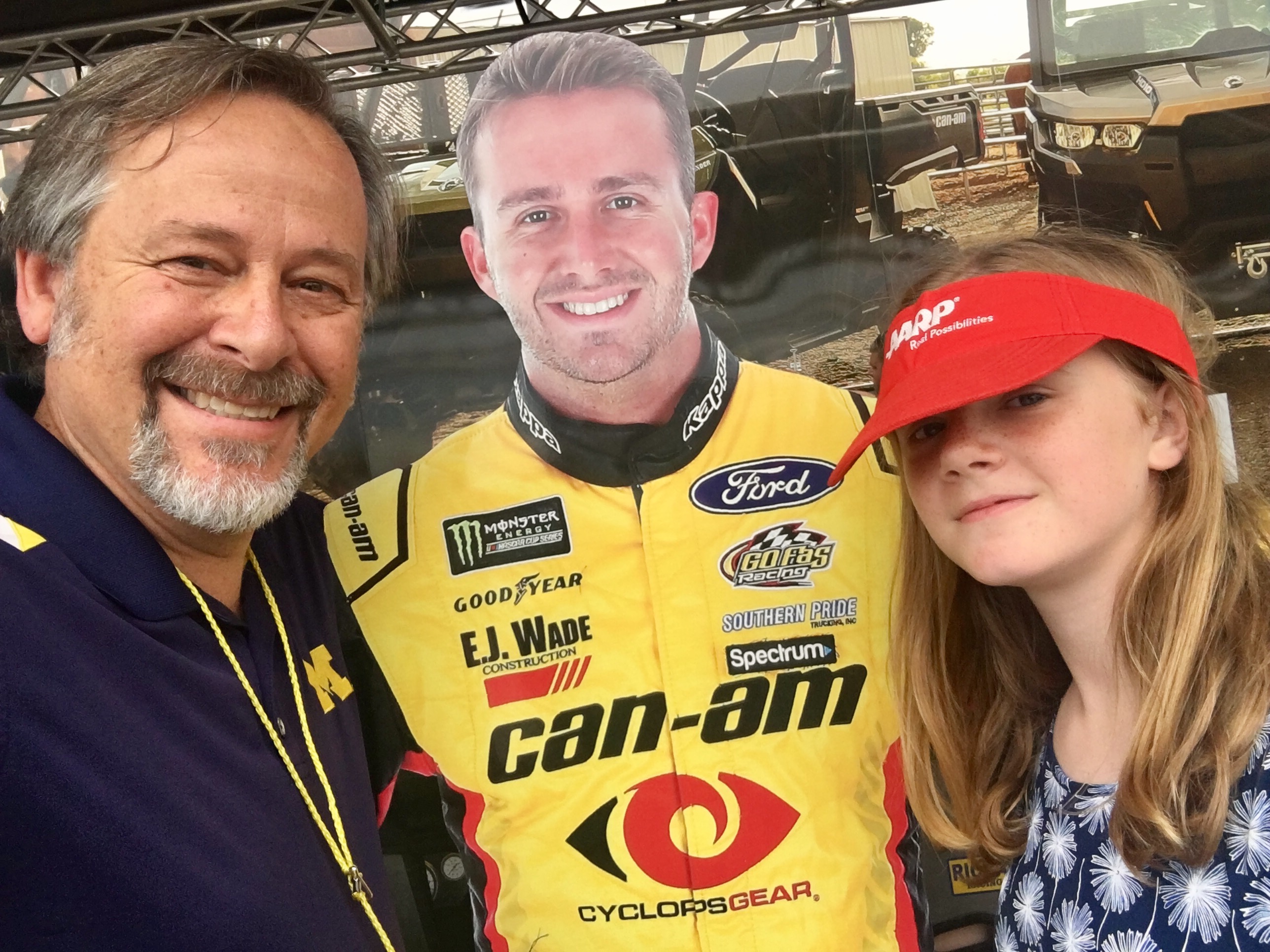 Man and girl with a red hat posing with cutout of nascar driver in yellow sponsor uniform