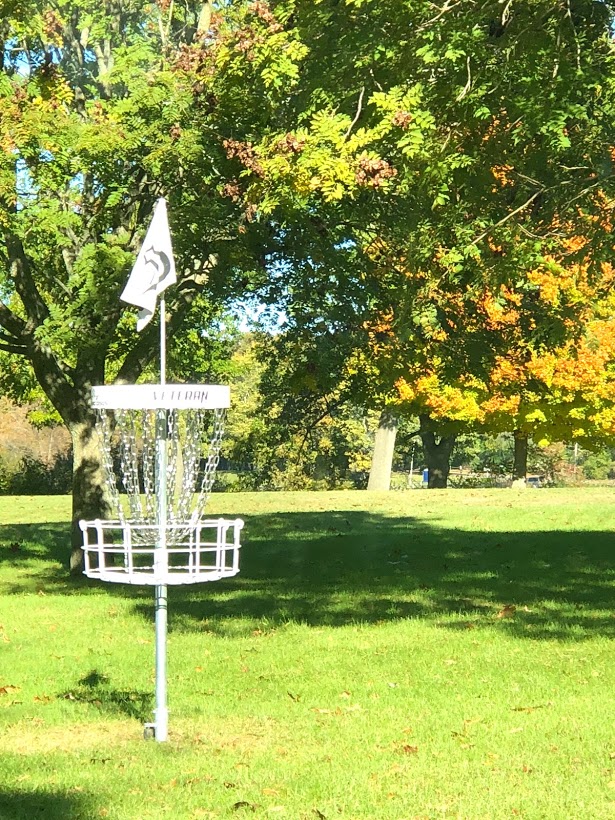 a white disc golf goal amidst green, yellow, and orange colored leaves during Fall colors season