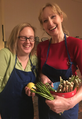 Tall blond woman red shirt short woman glasses green shirt blue aprons holding asparagus spears