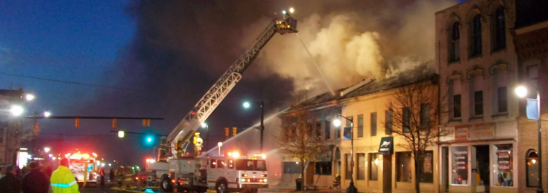 firefighter on ladder truck spraying water on burning building at night
